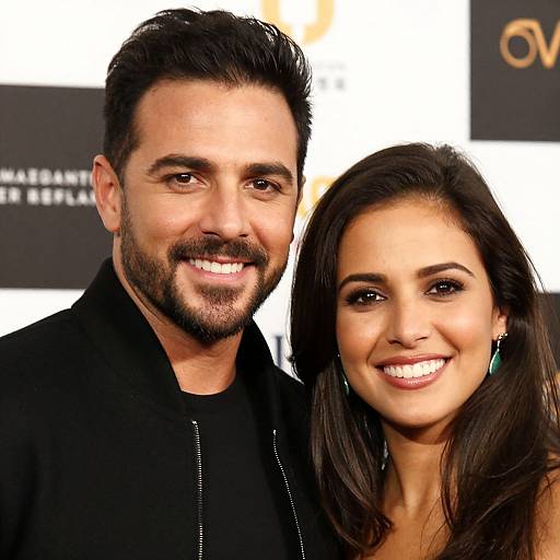 Photograph of a smiling couple; man with dark hair and beard, black shirt; woman with long brown hair, green earrings, black top. Background