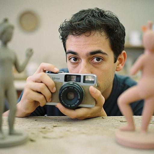 Photograph of a man with short black hair, intense focus, holding a vintage camera, photographing two small, blurred figurines on a table.