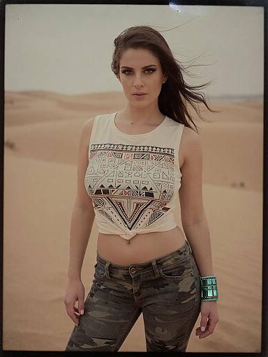Photograph of a confident woman with dark brown hair in a desert, wearing a white tribal-patterned crop top and camo jeans, with a turquoise