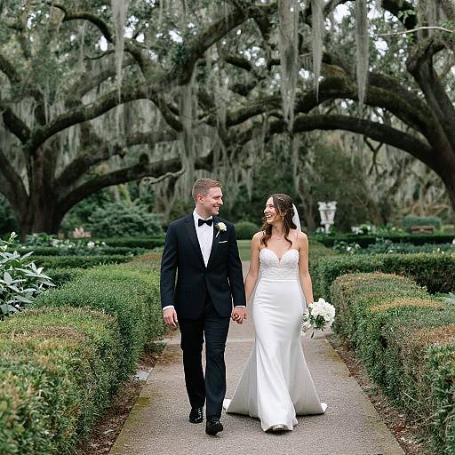 Photograph of a smiling bride in a strapless white gown and groom in a black tuxedo, holding hands and walking down a manicured garden