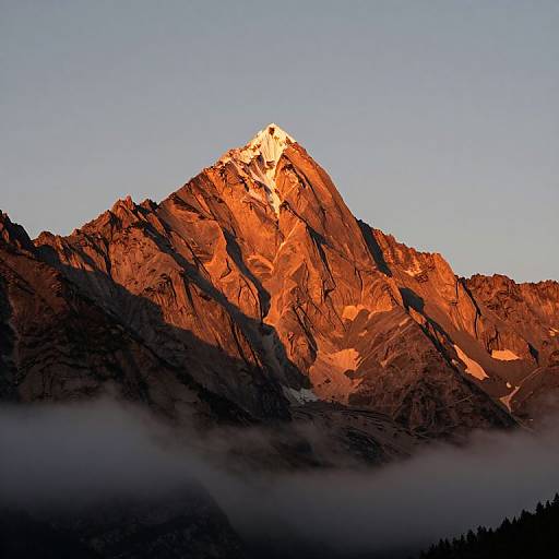 Photograph of a sunlit, rugged mountain peak with vibrant orange and red hues, contrasting against a clear blue sky and misty foreground.