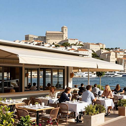 Photograph of a sunny Mediterranean waterfront restaurant with white umbrellas, patrons dining, and a historic town with a castle in the background.