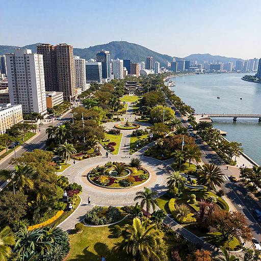 Aerial photograph of a sunlit coastal city park with palm trees, circular gardens, and high-rise buildings overlooking a blue harbor.