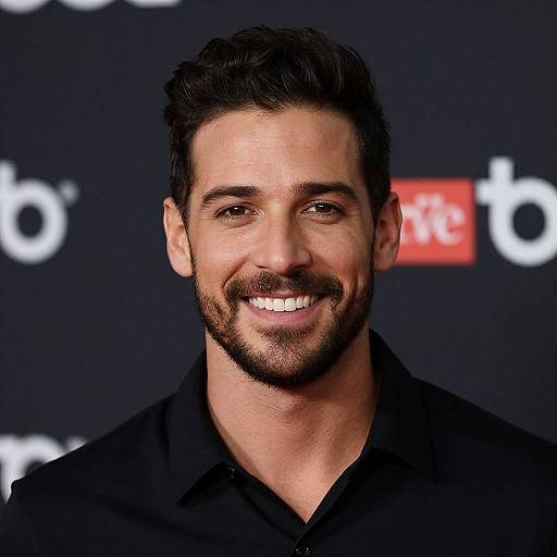 Photograph of a smiling, dark-haired, bearded man with a medium build, wearing a black shirt, against a blurred dark background with white and