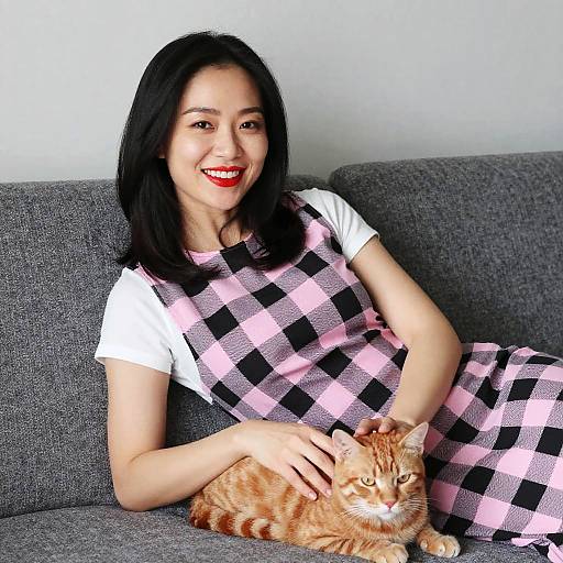 Photograph of an Asian woman with black hair, red lipstick, white shirt, pink and black checkered dress, smiling, sitting on a gray couch