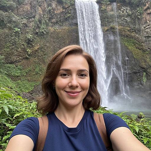 Photograph of a smiling young woman with medium-length brown hair, wearing a navy shirt and brown backpack, in front of a tall, lush waterfall.