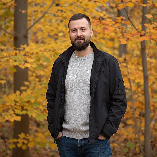 Photograph of a bearded man with short dark hair, wearing a black jacket and gray sweater, standing in an autumn forest with vibrant yellow and orange
