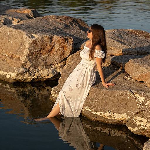 Photograph of a woman with long brown hair, wearing an off-shoulder white floral dress, sitting on rocky shoreline, legs in water, sunlight