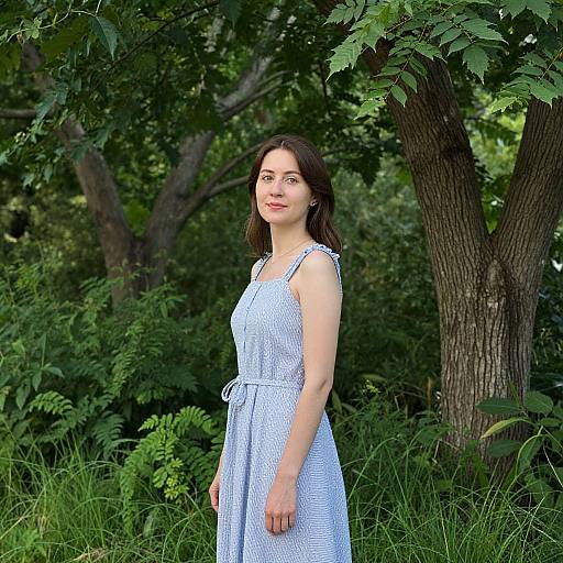 Photograph of a young woman with fair skin, brown hair, wearing a white, checkered sleeveless dress, standing in a lush green forest with
