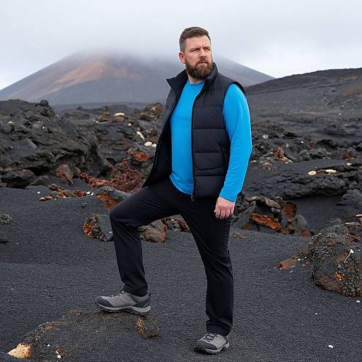 Photograph of a bearded man with short brown hair, wearing a blue shirt, black vest, and pants, standing confidently in a volcanic, rocky