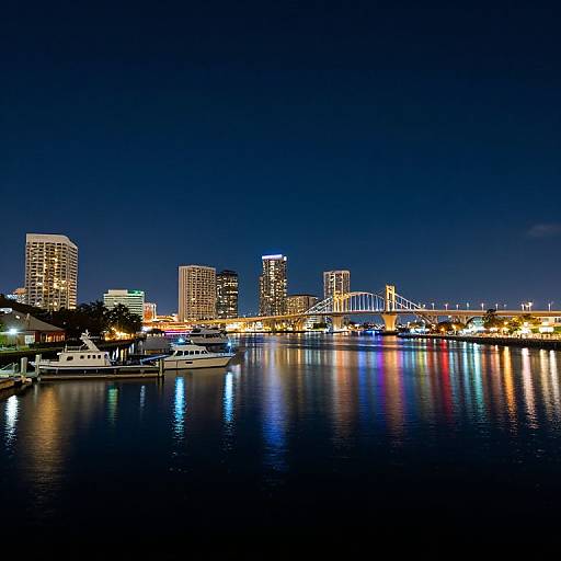 Photograph of a nighttime cityscape with illuminated skyscrapers, a brightly lit bridge, and colorful reflections on a calm river. Boats docked