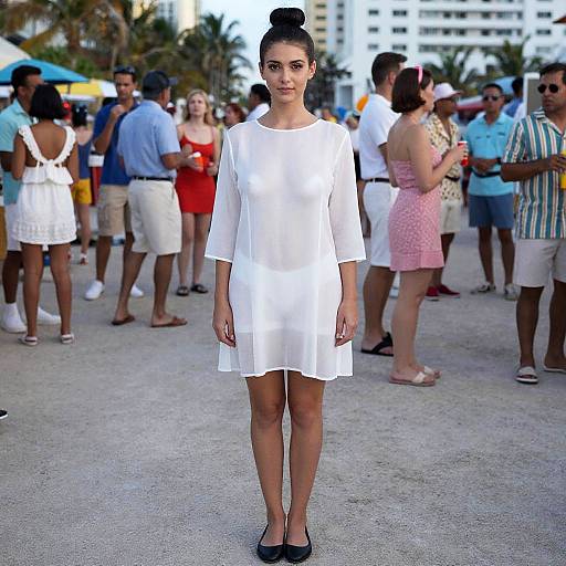 Photograph of a young woman with dark hair in a bun, wearing a sheer white dress and black flats, standing in front of a crowd at an