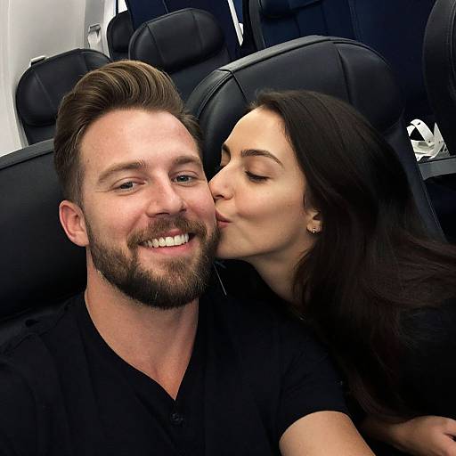Photograph of a smiling bearded man with brown hair, wearing a black shirt, as a woman with long dark hair kisses his cheek in a car