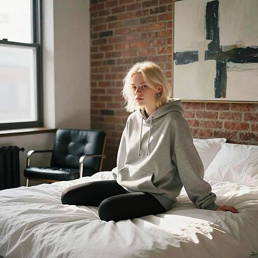 Photograph of a blonde woman in a gray hoodie and black pants, sitting on a sunlit bed against a brick wall with abstract art.