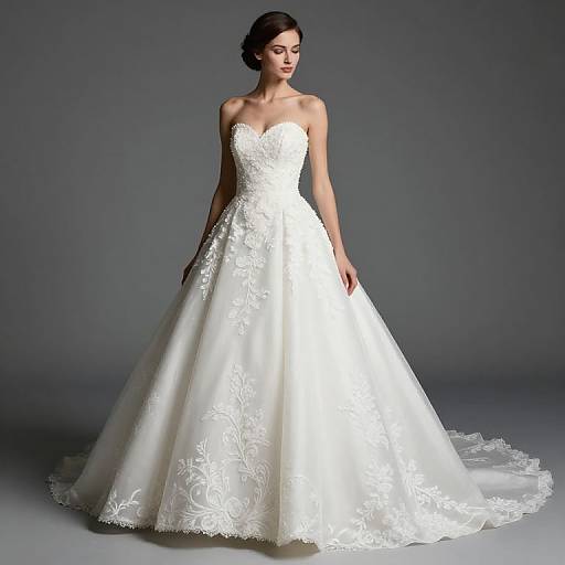 Photograph of a slender woman with dark hair in an elegant, strapless white wedding gown with lace and floral embroidery, standing against a gray background.