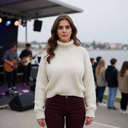 Photograph of a young woman with long brown hair, wearing a white knit turtleneck sweater and brown pants, standing outdoors in front of a music