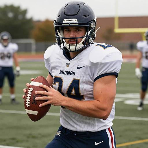 Photograph of a muscular male football player in white and navy uniform, gripping a football, wearing a black helmet, on a blurred field with other players