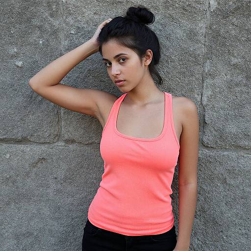 Photograph of a young woman with medium brown skin, black hair in a bun, wearing a neon pink tank top, black pants, standing against a