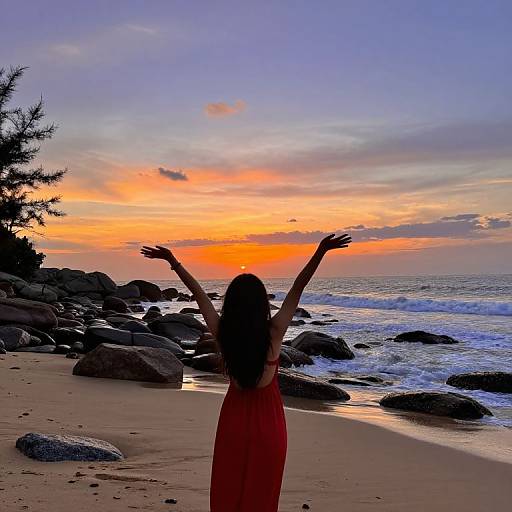 Photograph of a woman with long hair, arms raised, in a red dress, standing on a rocky beach at sunset with a colorful sky.