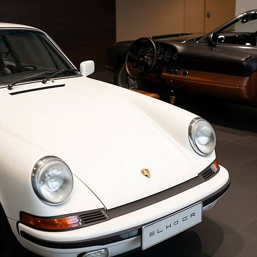 Photograph of a white classic Porsche 911 front view, headlights on, parked indoors beside a black Porsche, with a visible license plate.