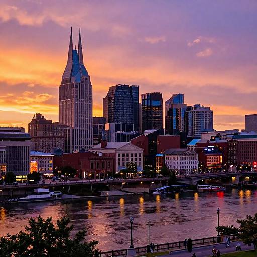 Photograph of a city skyline at sunset, featuring tall buildings with illuminated windows, a prominent twin-towered building, and a river reflecting vibrant orange,