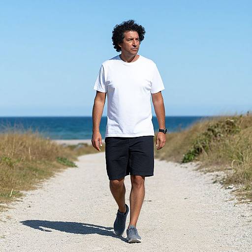 Photograph of a middle-aged man with curly black hair, wearing a white t-shirt, black shorts, and gray sneakers, walking on a sandy beach
