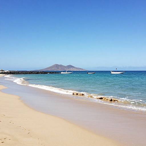 Photograph of a sunny beach with golden sand, blue ocean, small waves, distant rocky island, and three white boats floating.