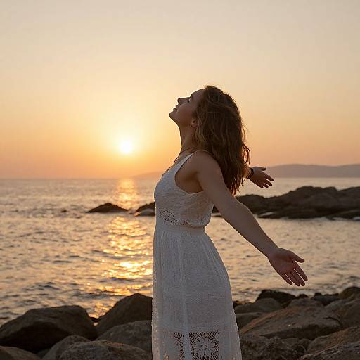 Photograph of a woman in a white lace dress, arms outstretched, head tilted back, enjoying a serene sunset over a rocky ocean shoreline.