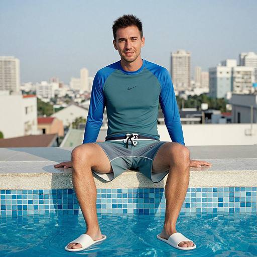 Photograph of a smiling, muscular man with short black hair, wearing a blue long-sleeve rash guard, gray shorts, and white sandals,