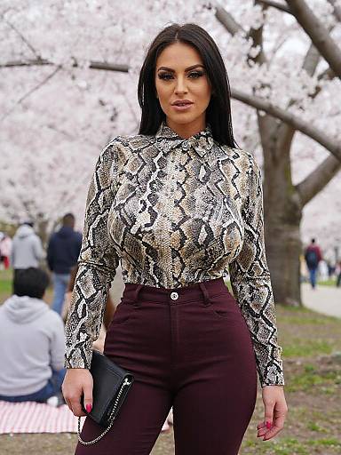 Photograph of a confident woman with long black hair, snake-print blouse, high-waisted maroon pants, black clutch, standing in a park