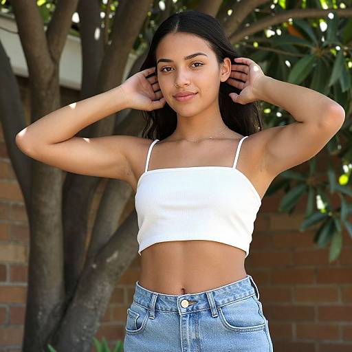 Photograph of a young woman with medium brown skin, long black hair, and slender build, wearing a white crop top and blue jeans, standing outdoors