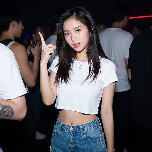 Photograph of an Asian woman with long black hair, wearing a white cropped t-shirt and blue jeans, pointing with her right index finger at a dim