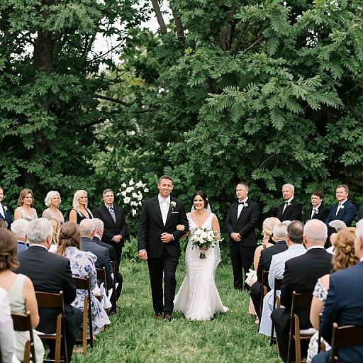 Photograph of a bride and groom walking down a grassy aisle, surrounded by seated guests in formal attire, with lush green trees in the background.