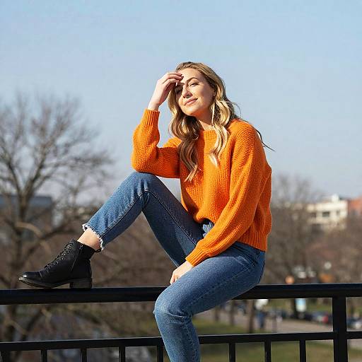 Photograph of a blonde woman in an orange sweater, blue jeans, and black boots, sitting on a black railing, smiling, with a sunny,