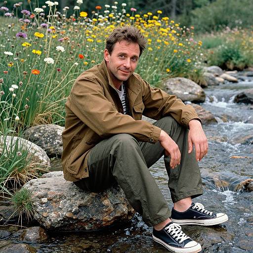 Photograph of a smiling middle-aged man with short brown hair, wearing a brown jacket, green pants, and black-and-white sneakers, sitting on a