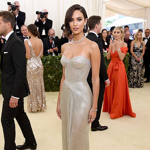 Photograph of a glamorous brunette woman in a sparkling, strapless silver gown, standing in a well-lit, elegant ballroom with photographers and attendees