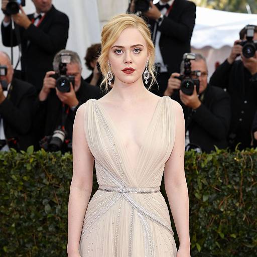 Photograph of a blonde woman with fair skin and red lipstick, wearing a sleeveless, white, pleated gown, standing in front of photographers and