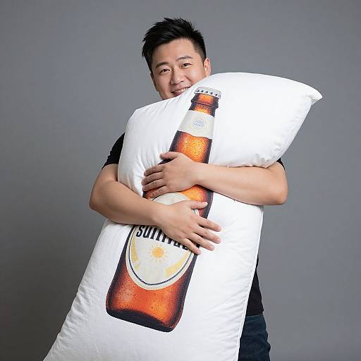 Photograph of an Asian man hugging a beer-themed white pillow, smiling, wearing a black shirt, against a plain gray background.