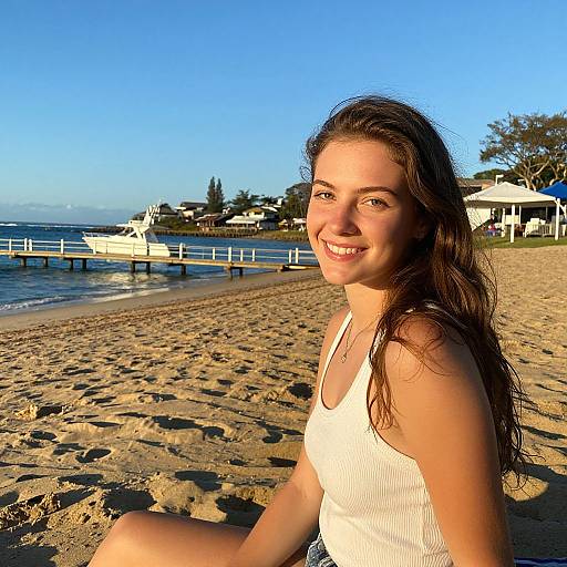Photograph of a smiling young woman with long brown hair, wearing a white tank top, sitting on a sandy beach at sunset, with a pier and