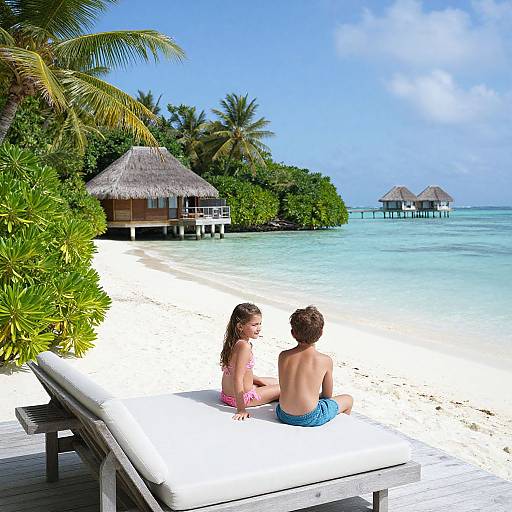 Photograph of a young couple sitting on a white-cushioned wooden bench on a tropical beach, overlooking overwater huts and calm turquoise water.