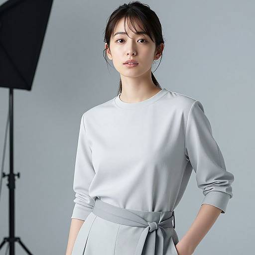 Photograph of an East Asian woman with fair skin, black hair in a ponytail, wearing a light gray, long-sleeve dress, standing