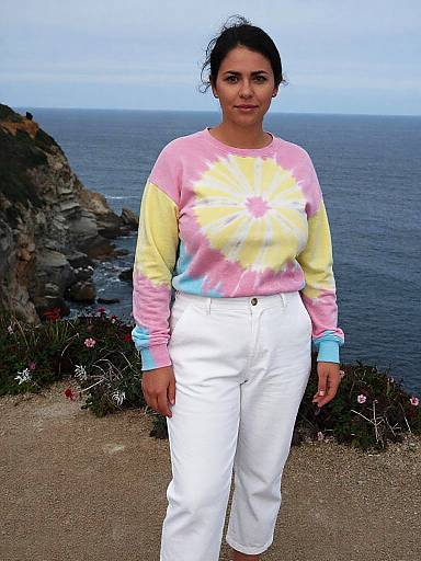 Photograph of a woman with medium skin tone, dark hair, wearing a pastel tie-dye sweater and white pants, standing by a rocky coastal