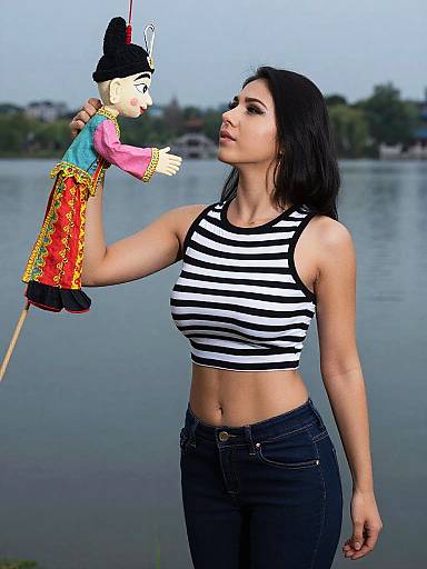 Photograph of a young woman with black hair, wearing a black and white striped crop top and dark jeans, holding a colorful, traditional puppet against a