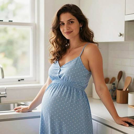 Photograph of a pregnant woman with long brown hair, wearing a light blue polka dot sundress, standing in a bright, modern kitchen.