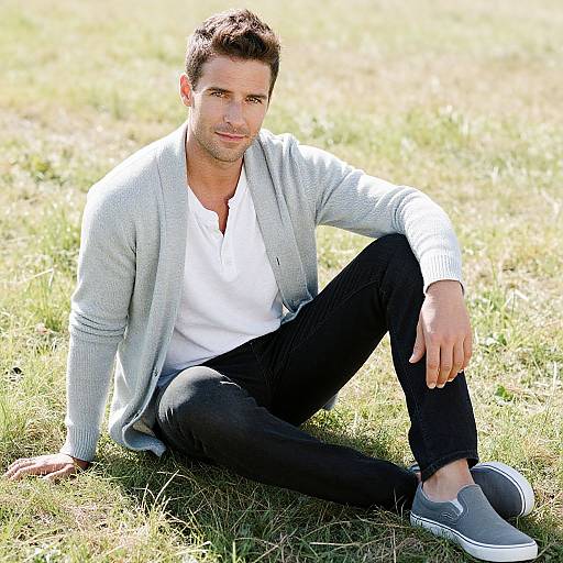 Photograph of a handsome, dark-haired man with light skin, sitting on grass, wearing a white shirt, gray cardigan, black pants, and
