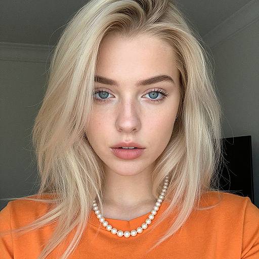 Photograph of a blonde woman with blue eyes, wearing an orange shirt and white pearl necklace, looking directly at the camera.