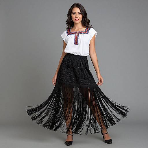 Photograph of a dark-haired woman in a white blouse with red embroidery, black fringe skirt, and black heels, standing against a gray background.