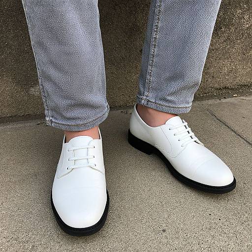 Photograph of white leather loafers with black soles on light blue, slightly frayed denim jeans, standing on concrete pavement.