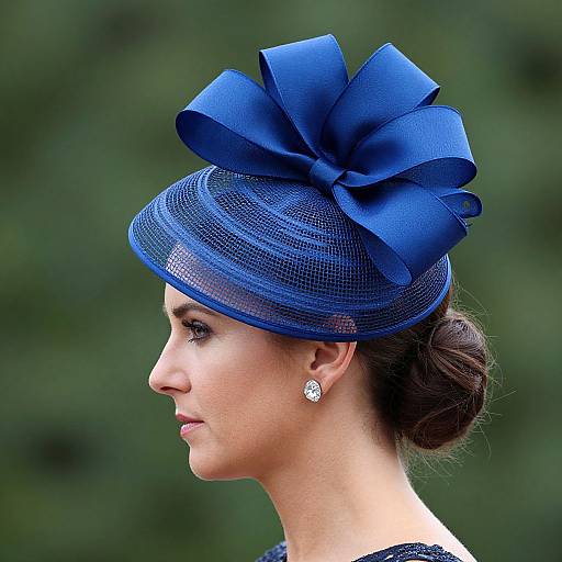 Photograph of a woman in profile, wearing a vibrant blue, mesh fascinator with a large ribbon bow, and sparkling earrings, against a blurred green