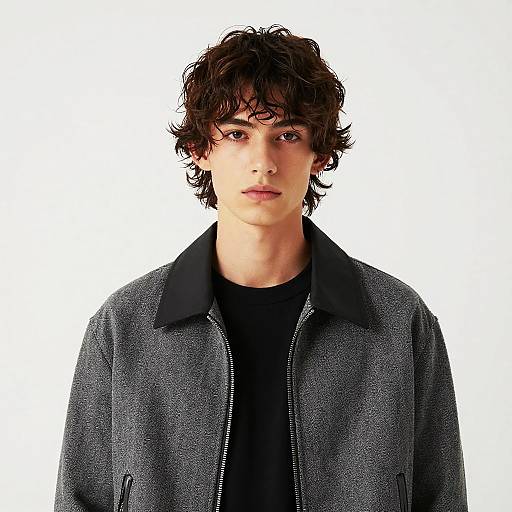Photograph of a young man with curly brown hair, wearing a black collared shirt and gray textured jacket, against a plain white background.